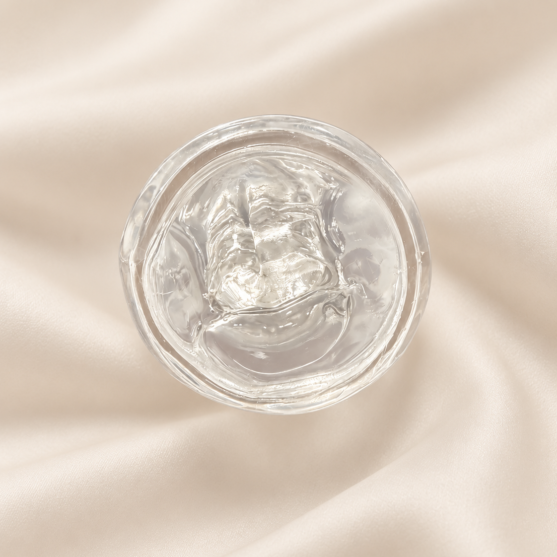 A clear, gel-like cosmetic product in a round transparent container, photographed from above on a softly draped cream satin fabric background.