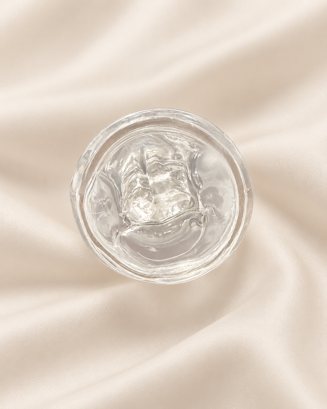 A clear, gel-like cosmetic product in a round transparent container, photographed from above on a softly draped cream satin fabric background.