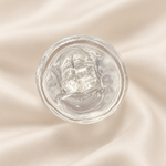A clear, gel-like cosmetic product in a round transparent container, photographed from above on a softly draped cream satin fabric background.
