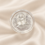 A clear, gel-like cosmetic product in a round transparent container, photographed from above on a softly draped cream satin fabric background.